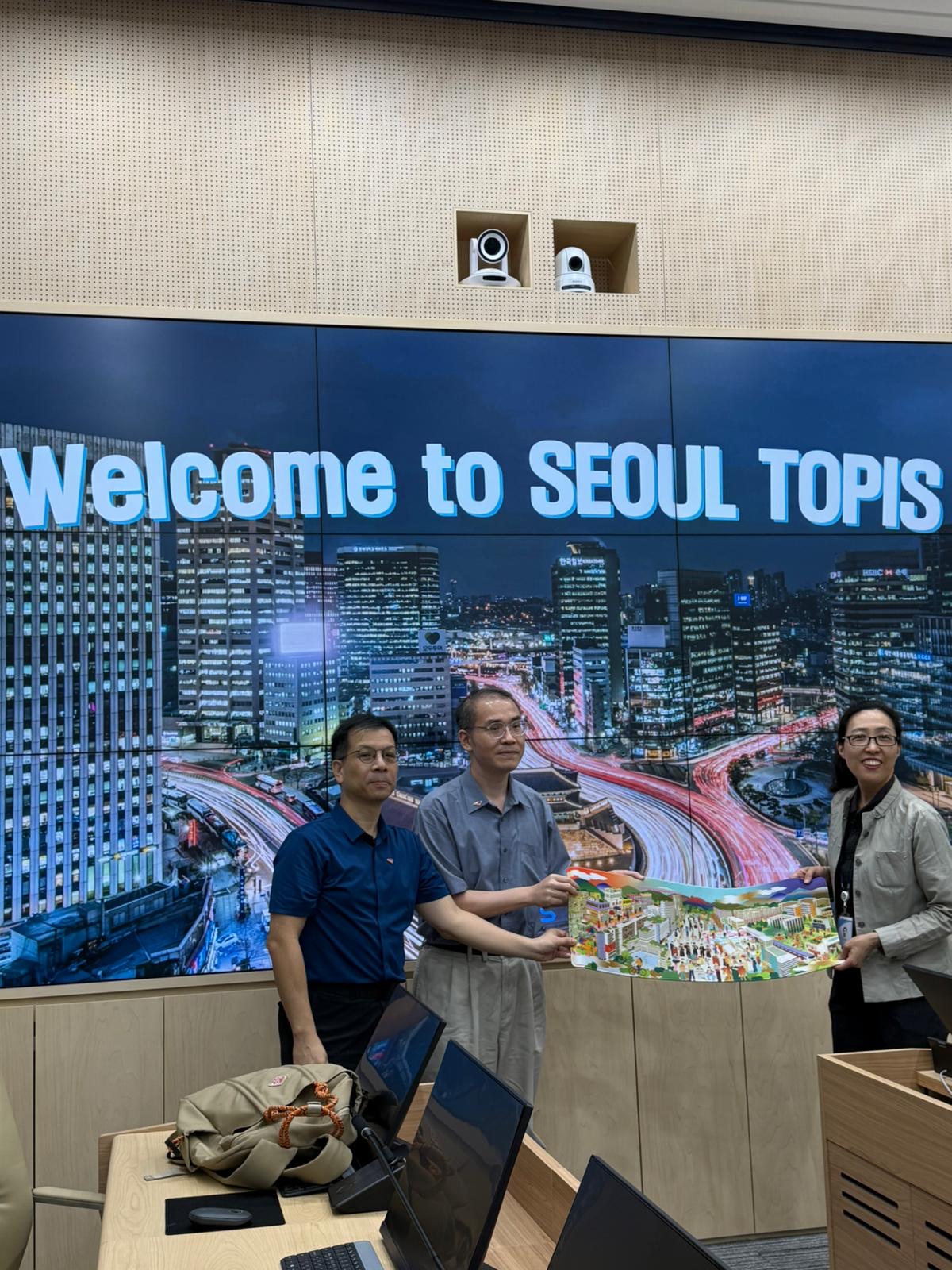 A souvenir exchange in front of the glowing Welcome to SEOUL TOPIS cityscape screen